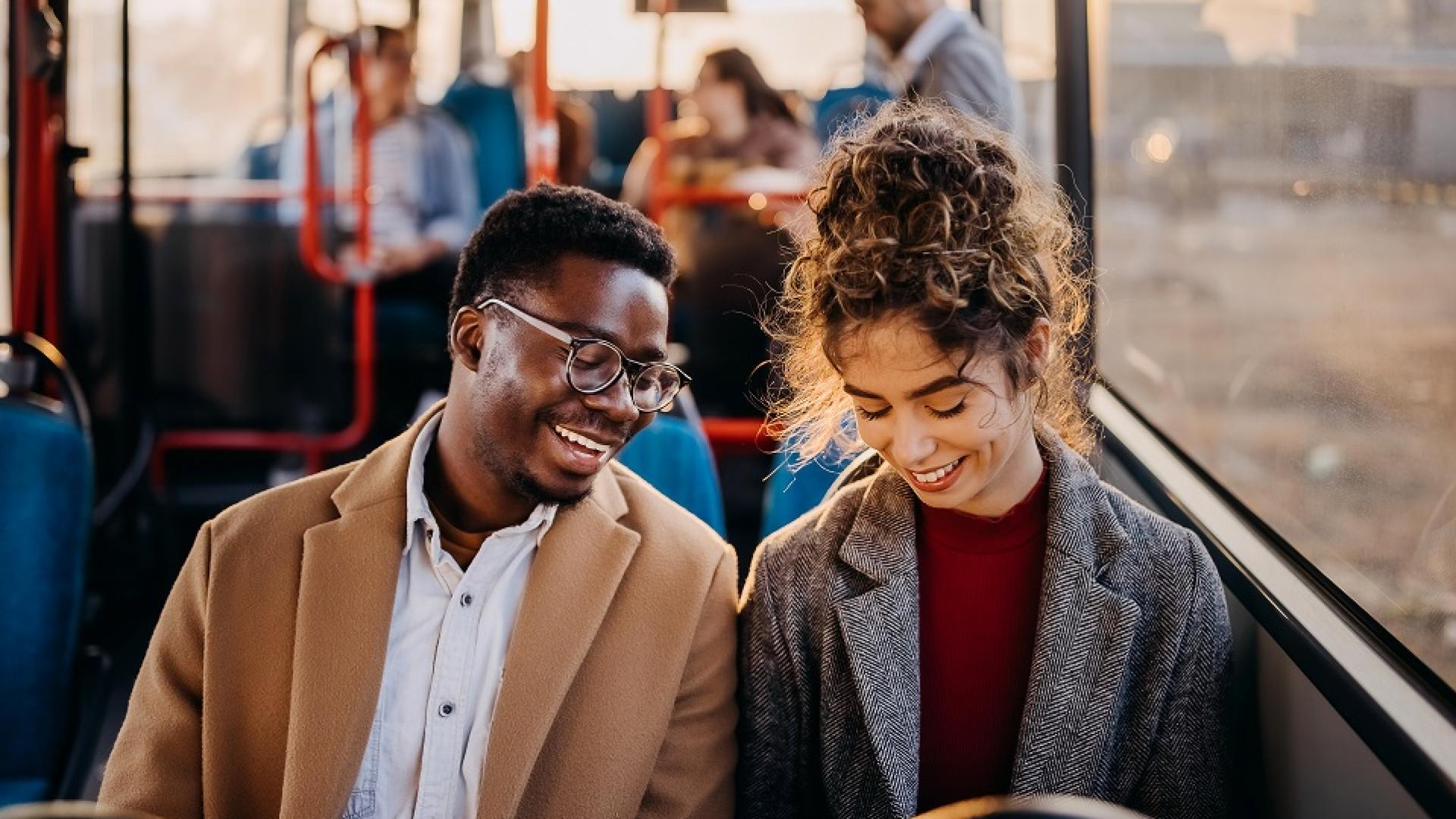 jeune homme noir avec des lunette et jeune femme blanche avec un chignon souriants assis passagers d'un bus