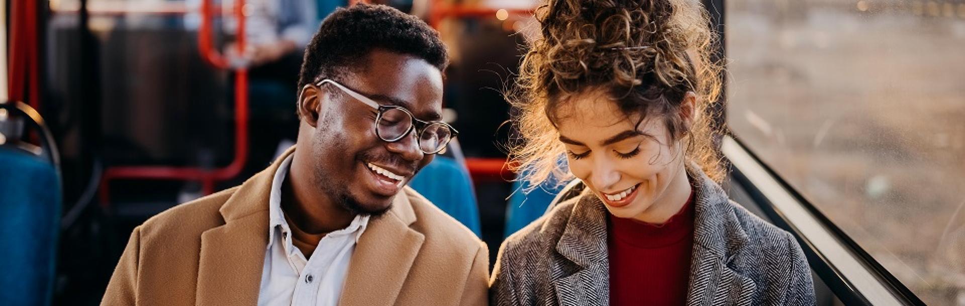 jeune homme noir avec des lunette et jeune femme blanche avec un chignon souriants assis passagers d'un bus