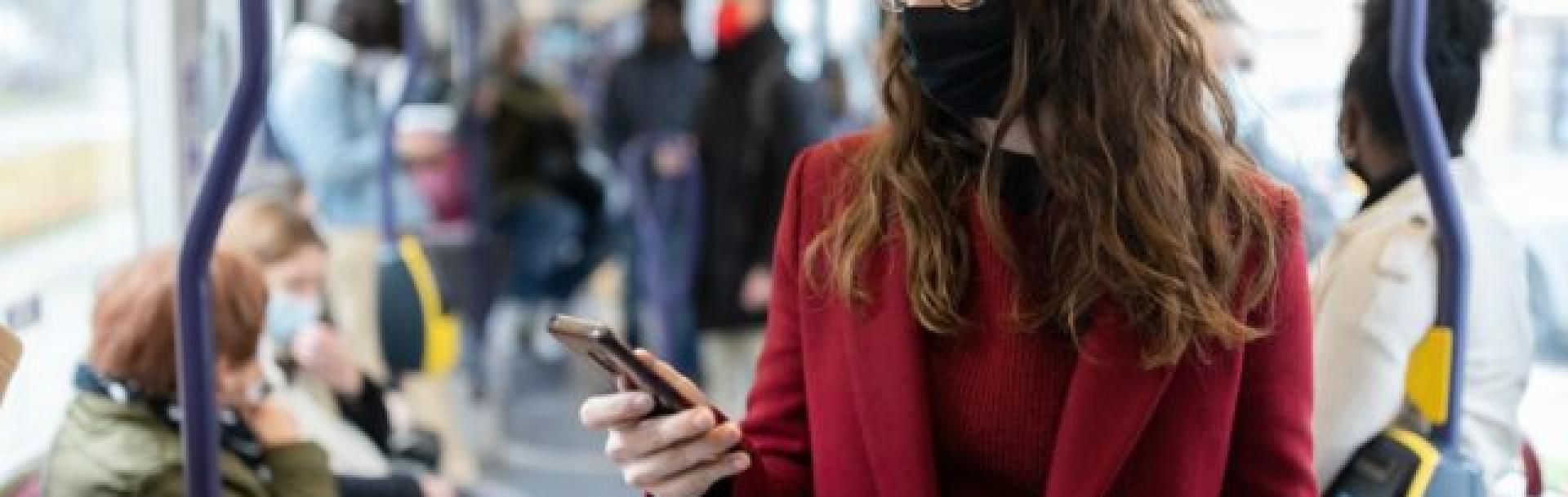 Femme masque noir manteau et pull rouge avec lunette dans bus bleu regardant son téléphone portable