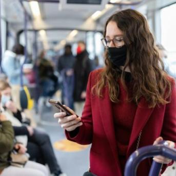 Femme masque noir manteau et pull rouge avec lunette dans bus bleu regardant son téléphone portable