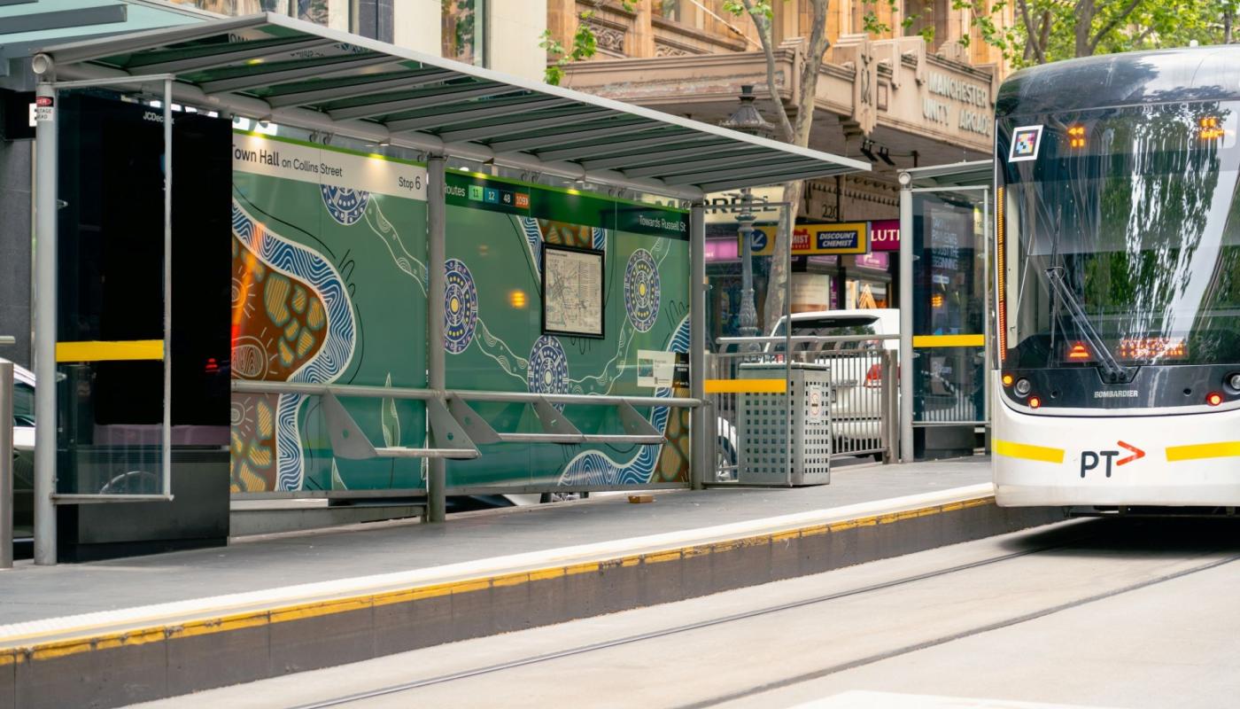 “Town Hall” tram shelter on Collins Street in Melbourne, featuring a First Nations artwork (Aboriginal motifs) as part of Yarra Trams' Art Stops initiative. Alt EN: “Town Hall” tram stop on Collins Street, Melbourne, wrapped in First Nations