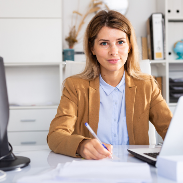 woman writing on her desk with her computer in brown jacket and blue shirt