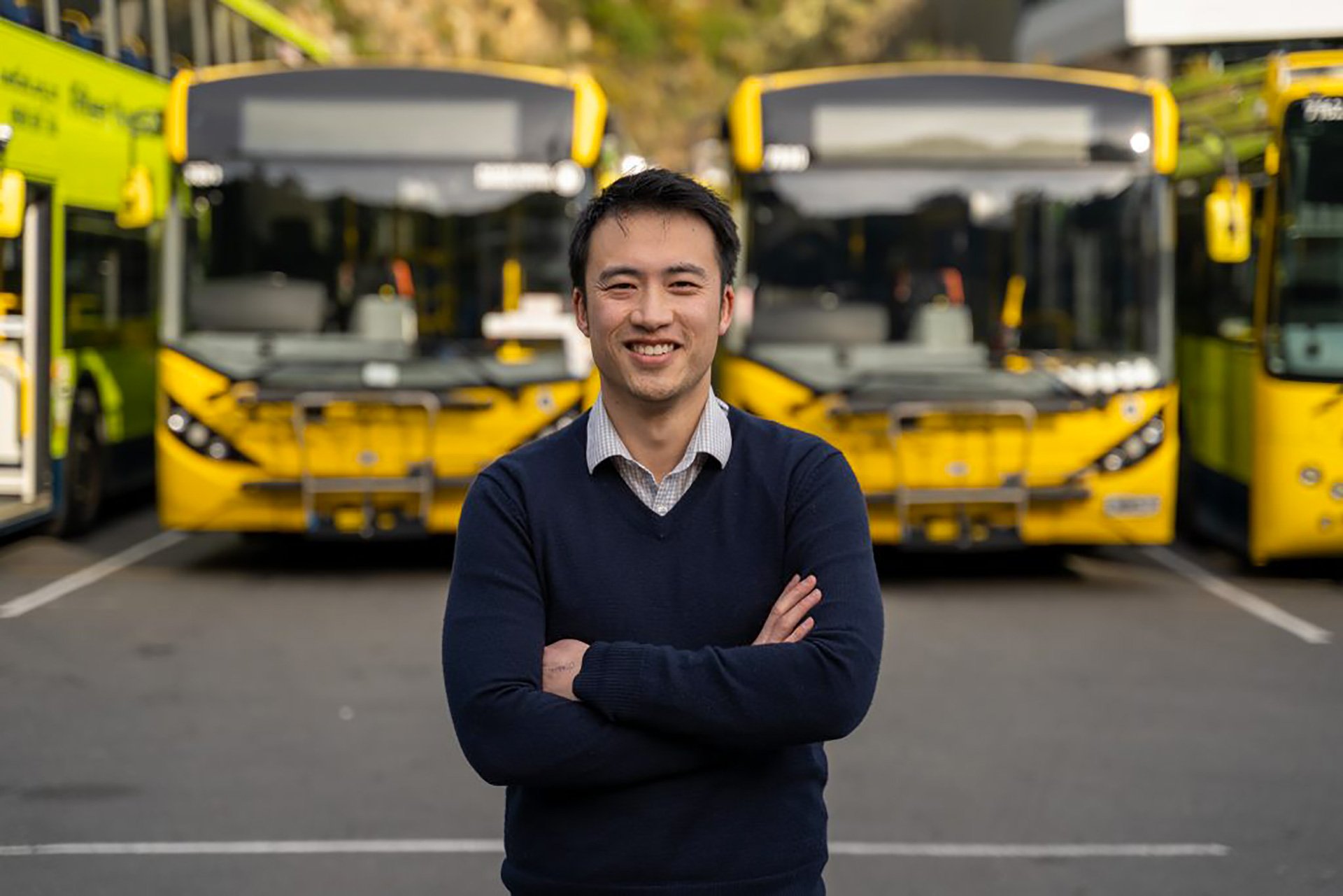 smiling man with crossed arms and blue sweater blurred background with yellow bus