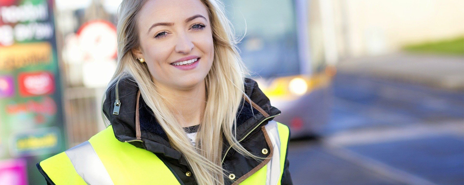 jeune femme blonde souriante avec un gilet de sécurité jaune fluorescent posant devant un bus, l'arrière-plan est flou