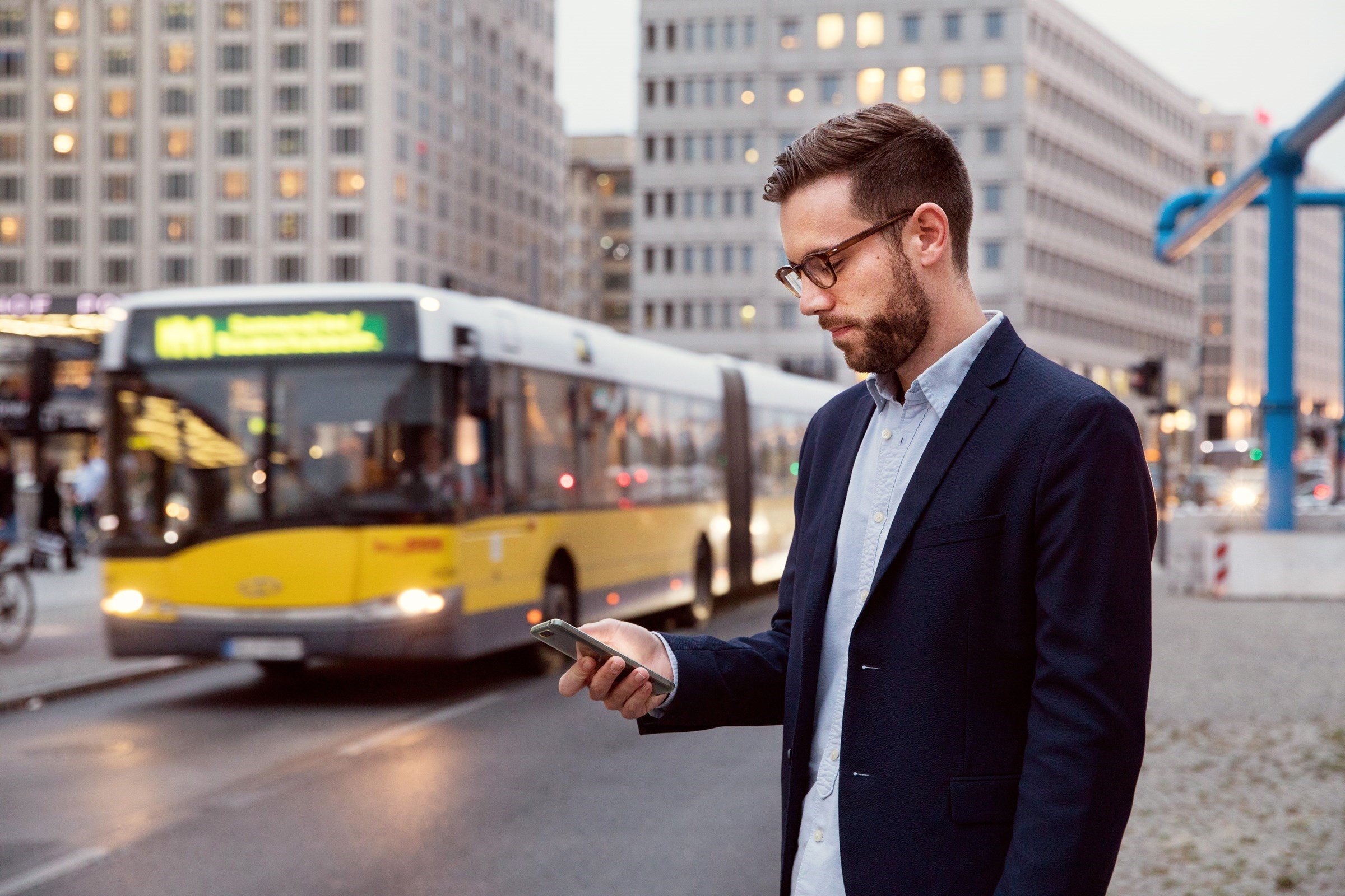 man in blue jacket looking at his smartphone blurred background with yellow bus