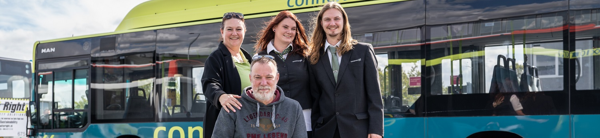 man on a wheelchair with three people behind in black uniform smiling in front of a pavement and a blue and green bus connection in the background