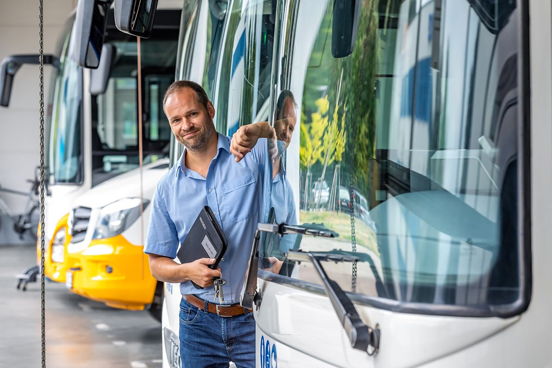 man leaning on a white and yellow bus in a blue shirt and jeans with a file in his hand