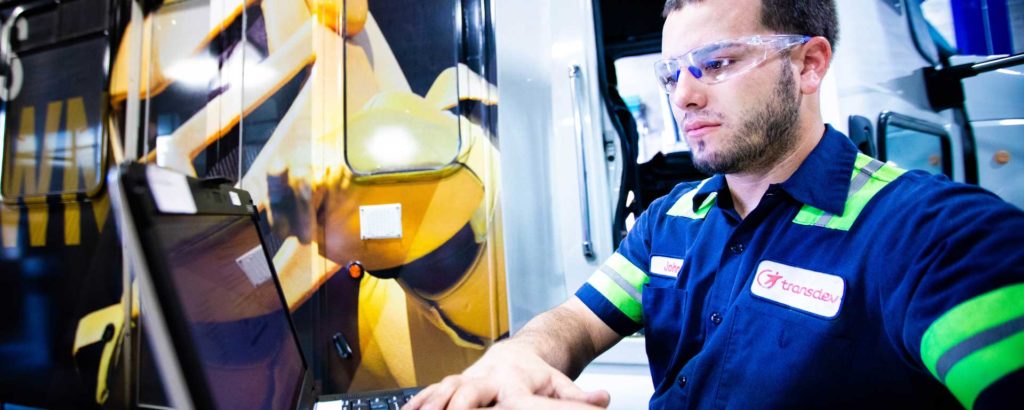 homme en uniforme de travail vert et bleu avec lunette de protection travaillant sur un ordinateur devant un camion