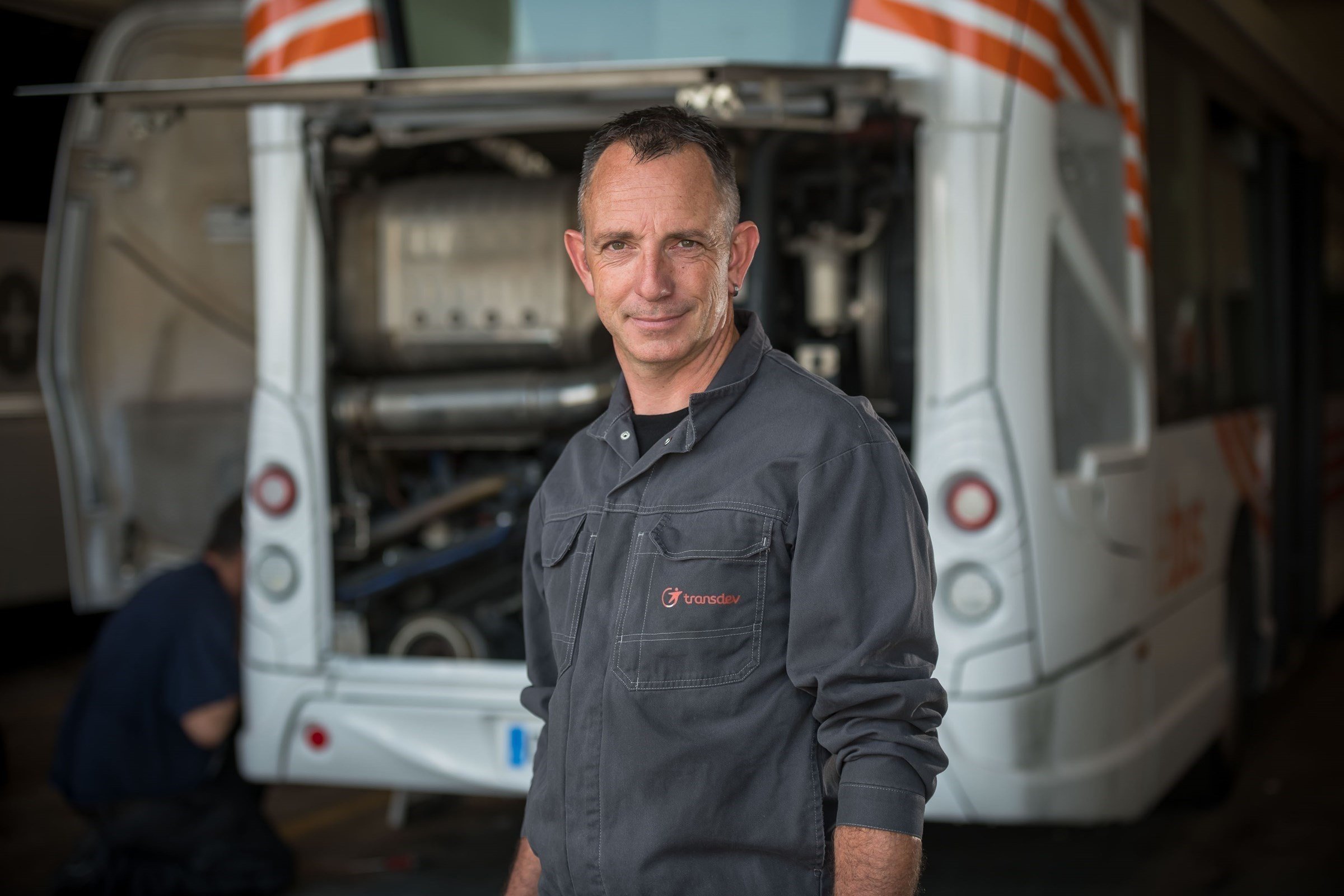 smiling man in a dark grey shirt, posing in front of an open trunk of a bus