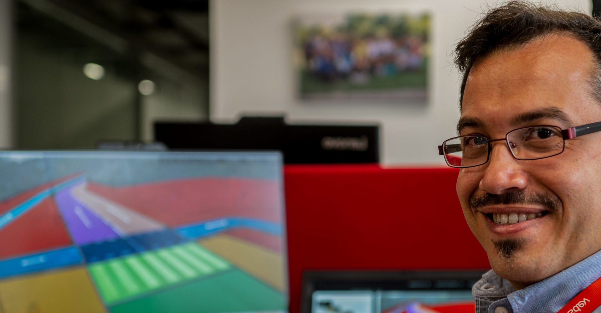 man wearing glasses looking and smiling at the camera with a background in a blurred office