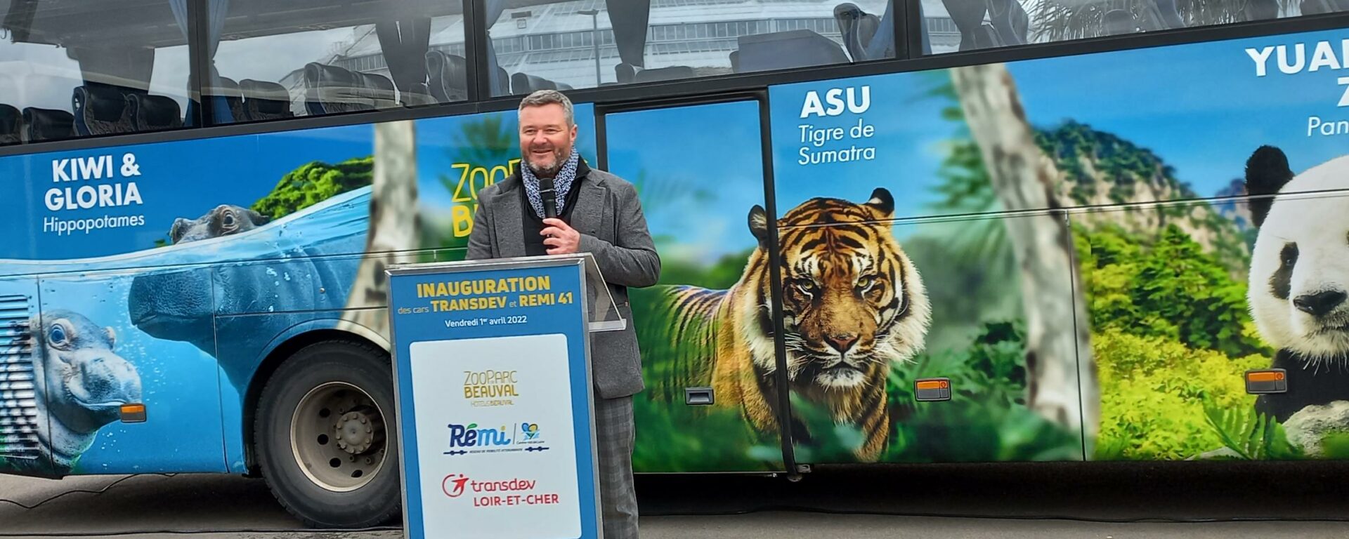 Un homme debout à une tribune, micro à la main, devant un car au covering Zoo de Beauval