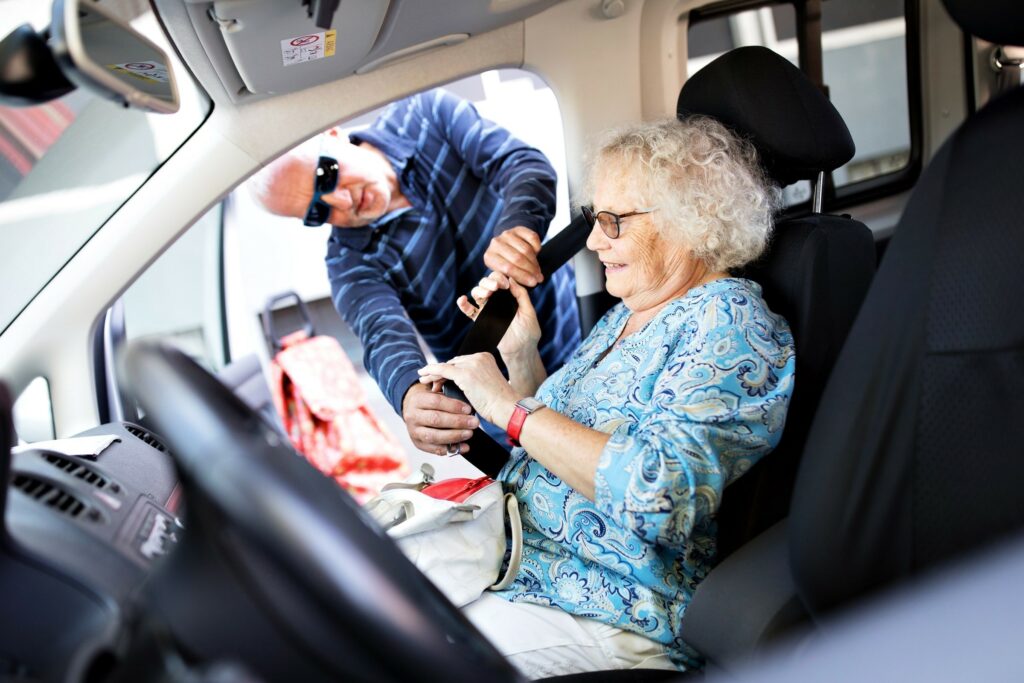 Un homme debout à la portière d'un véhicule attache la ceinture de sécurité d'un dame âgée.