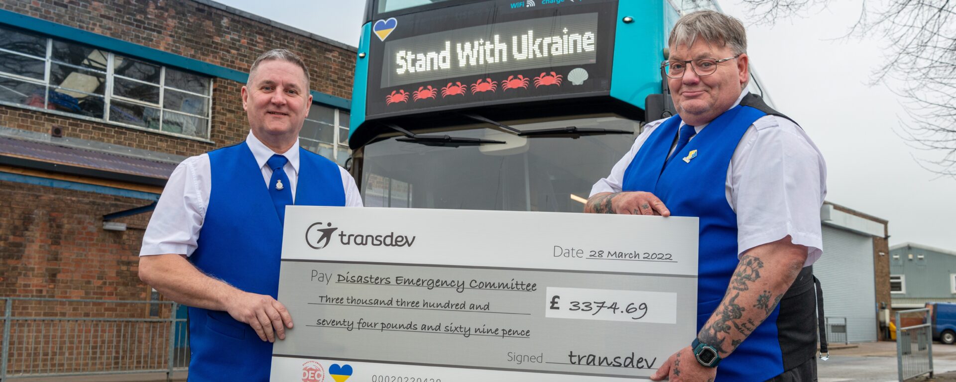 2 employés de Transdev UK en uniforme se tiennent debout devant un bus à 2 étages bleu, et brandissent un chèque géant avec de montant des sommes récoltées pour soutenir l'Ukraine