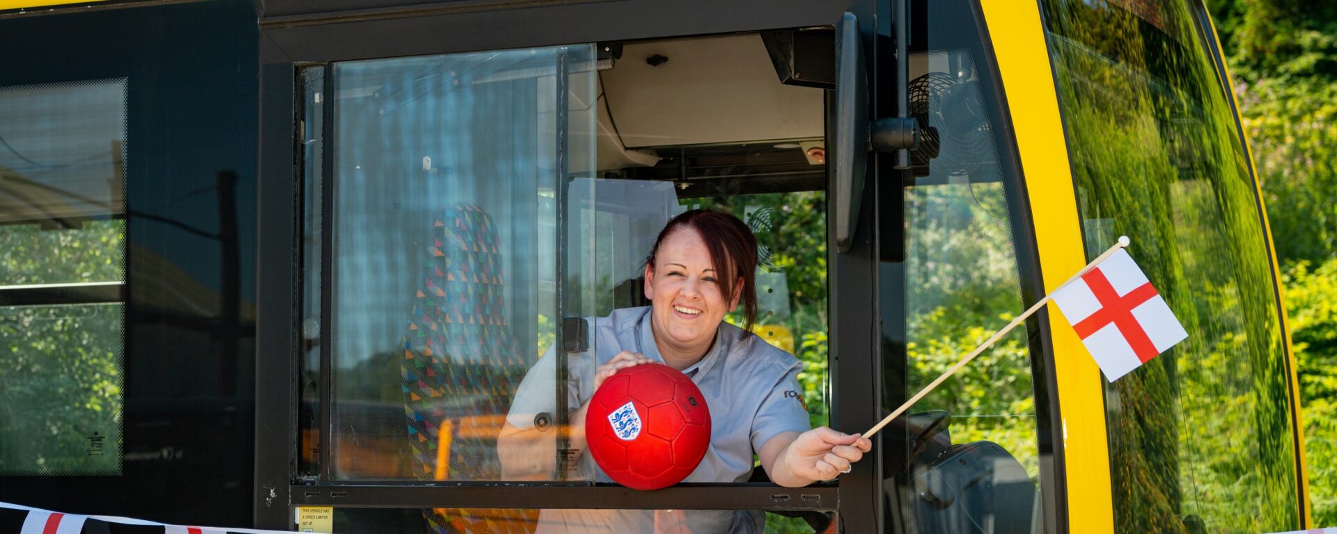 Conductrice de bus penchée à la fenêtre d'un bus jaune qui porte le nom de la joueuse Keira Walsh. Elle agite un fanion aux couleurs de l'Angleterre, et tient un ballon de foot aux couleurs de l'Angleterre dans l'autre main