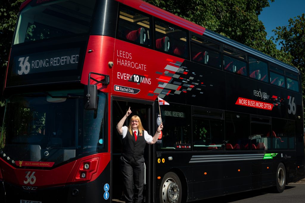 One Leeds Harrogate female bus driver stands at the entrance of the bus named after Rachel Daily, holding an England red and white flag